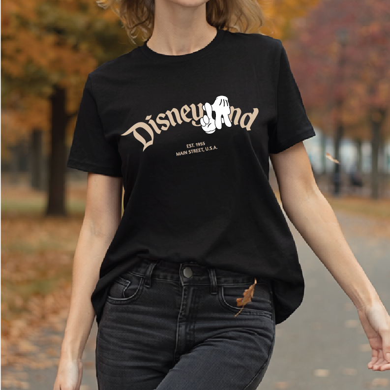 A woman wearing a black short-sleeve graphic T-shirt with a white 'Disneyland' script and Mickey Mouse silhouette, paired with dark jeans, photographed outdoors on a path with autumn trees in the background.