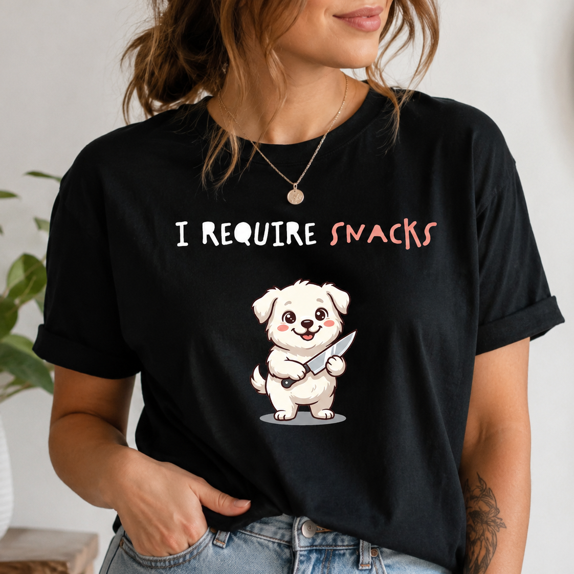 A woman wearing a black graphic t-shirt with the text 'I REQUIRE SNACKS' and a cartoon dog holding a snack. The photo is taken in a lifestyle setting with a potted plant in the background.