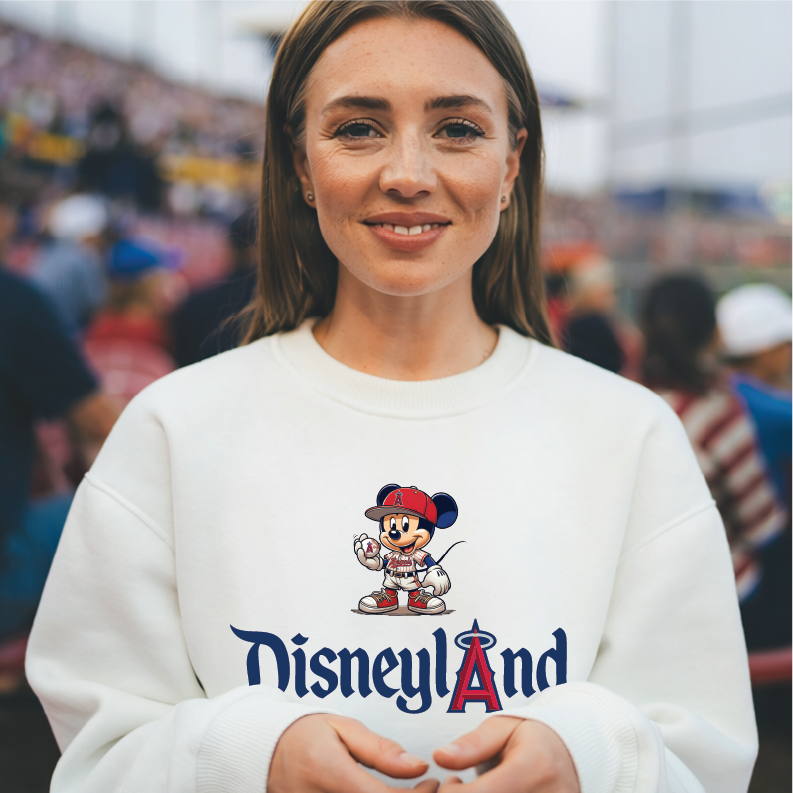 A woman wearing a white crewneck sweatshirt with a Mickey Mouse baseball graphic and the Disneyland logo, photographed in a crowded outdoor stadium setting.