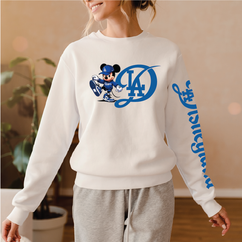 A woman wearing a white crewneck sweatshirt with a blue Los Angeles Dodgers Mickey Mouse graphic on the chest and blue script text down the right sleeve, paired with gray sweatpants in a warm indoor setting.