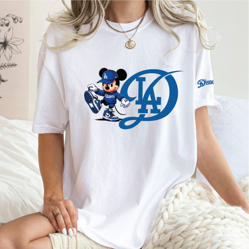 A woman wearing a white short-sleeve t-shirt with a blue Los Angeles Dodgers Mickey Mouse graphic on the front, sitting on a bed with a chunky knit blanket.