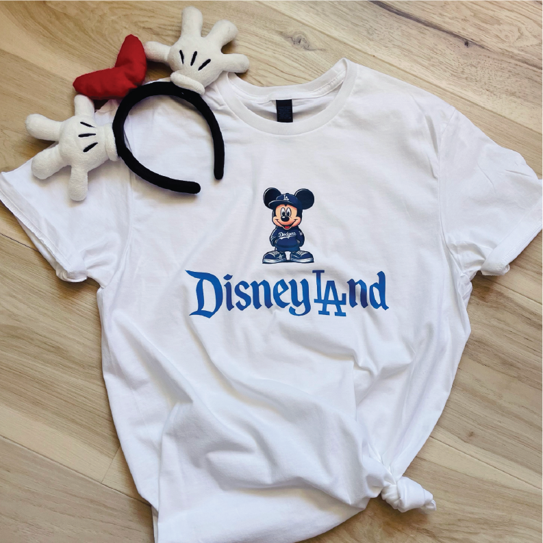A white short-sleeve T-shirt laid flat on a light wood floor, featuring a blue 'Disneyland' logo and Mickey Mouse graphic on the chest. A plush Minnie Mouse headband accessory with a red bow and white gloves is placed on top of the shirt.