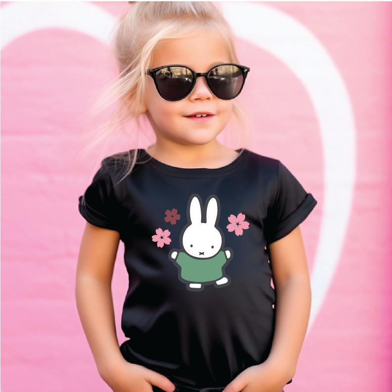 A young girl with blonde hair wearing black sunglasses and a black short-sleeve t-shirt with aMiffy graphic, standing against a pink background with a white heart outline.