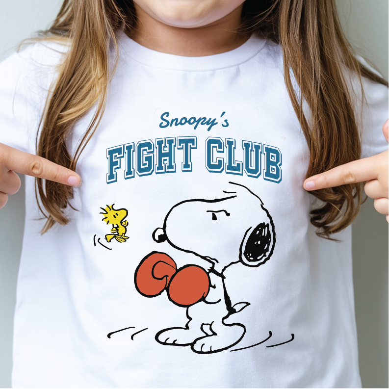 A child wearing a white graphic T-shirt with Snoopy and Woodstock artwork and the text 'Snoopy's FIGHT CLUB,' with both hands pointing at the shirt.