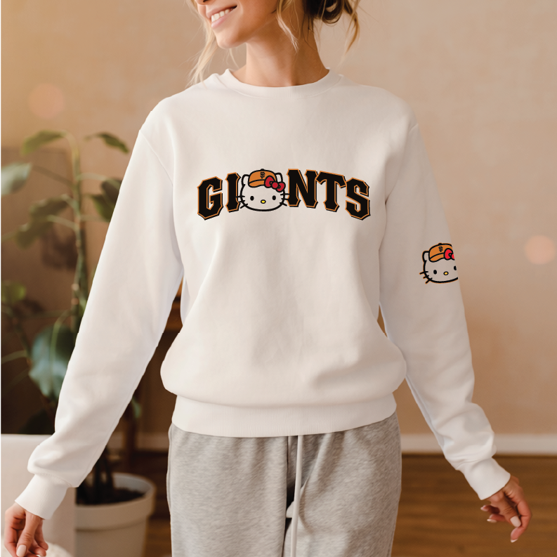 A woman wearing a white crewneck sweatshirt with 'GIANTS' and a Hello Kitty face graphic across the chest, paired with gray sweatpants in a warm indoor setting.