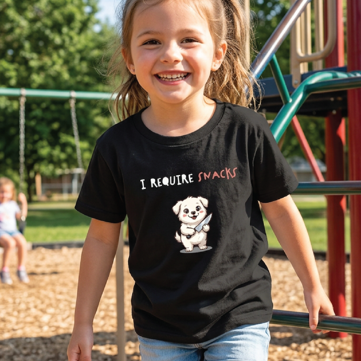 A young girl smiling on a playground wearing a black graphic t-shirt that reads 'I REQUIRE SNACKS' with a cartoon puppy holding a bag of chips.
