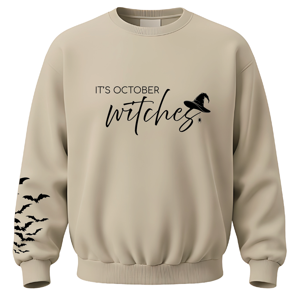 A beige crewneck sweatshirt with a black graphic print on the front reading 'IT'S OCTOBER witches' alongside a witch hat silhouette, and black bat silhouettes printed along the right sleeve.