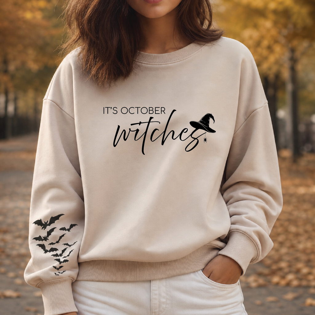 A woman wearing a beige crewneck sweatshirt with a black graphic that reads 'IT'S OCTOBER witches' and a witch hat, with bat prints on the sleeve, set against an autumn park background.