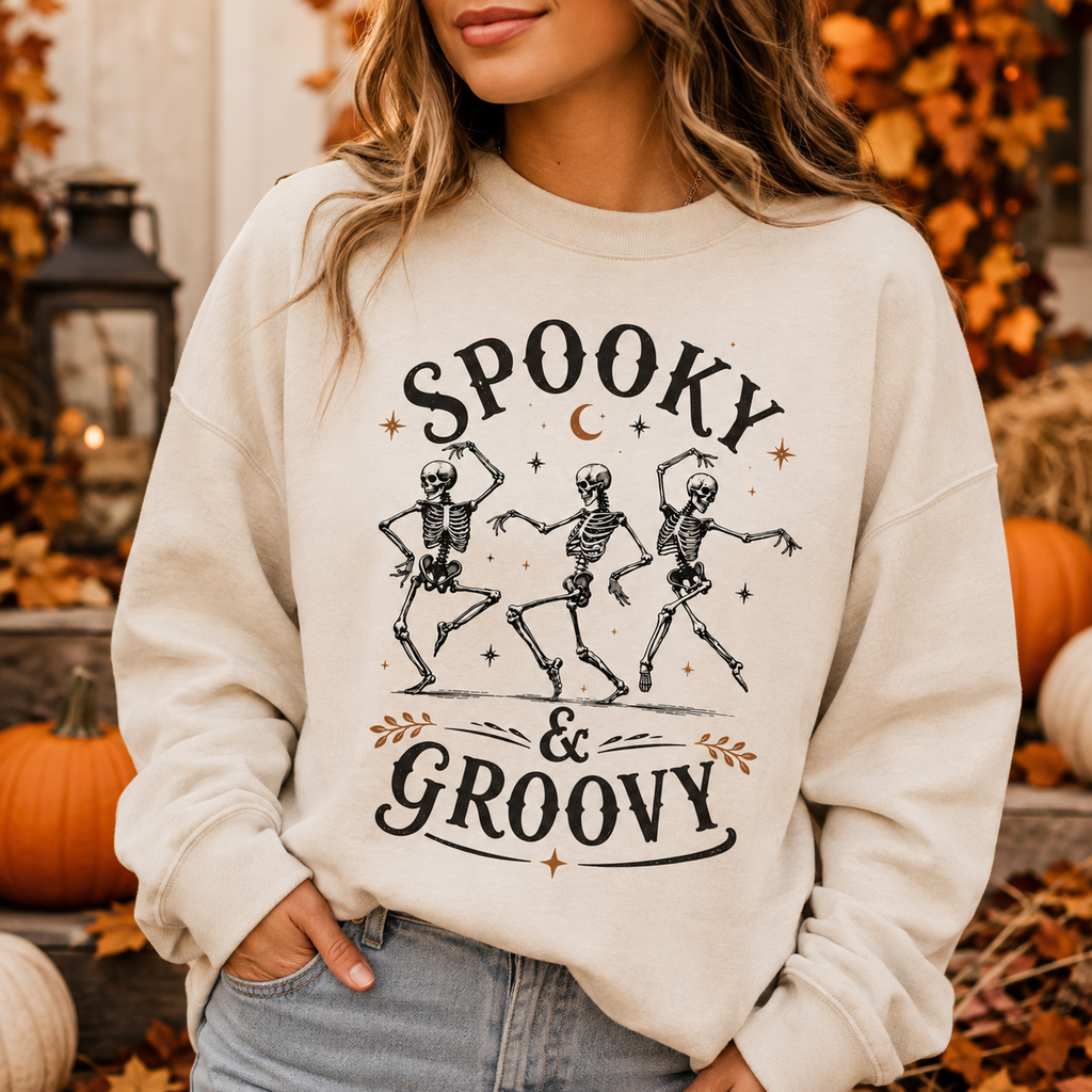 A woman wearing a cream-colored crewneck sweatshirt with a black and orange graphic that reads 'SPOOKY & GROOVY' with three dancing skeletons. The background is a blurred autumn-themed outdoor setting with pumpkins, fall leaves, and a lantern.