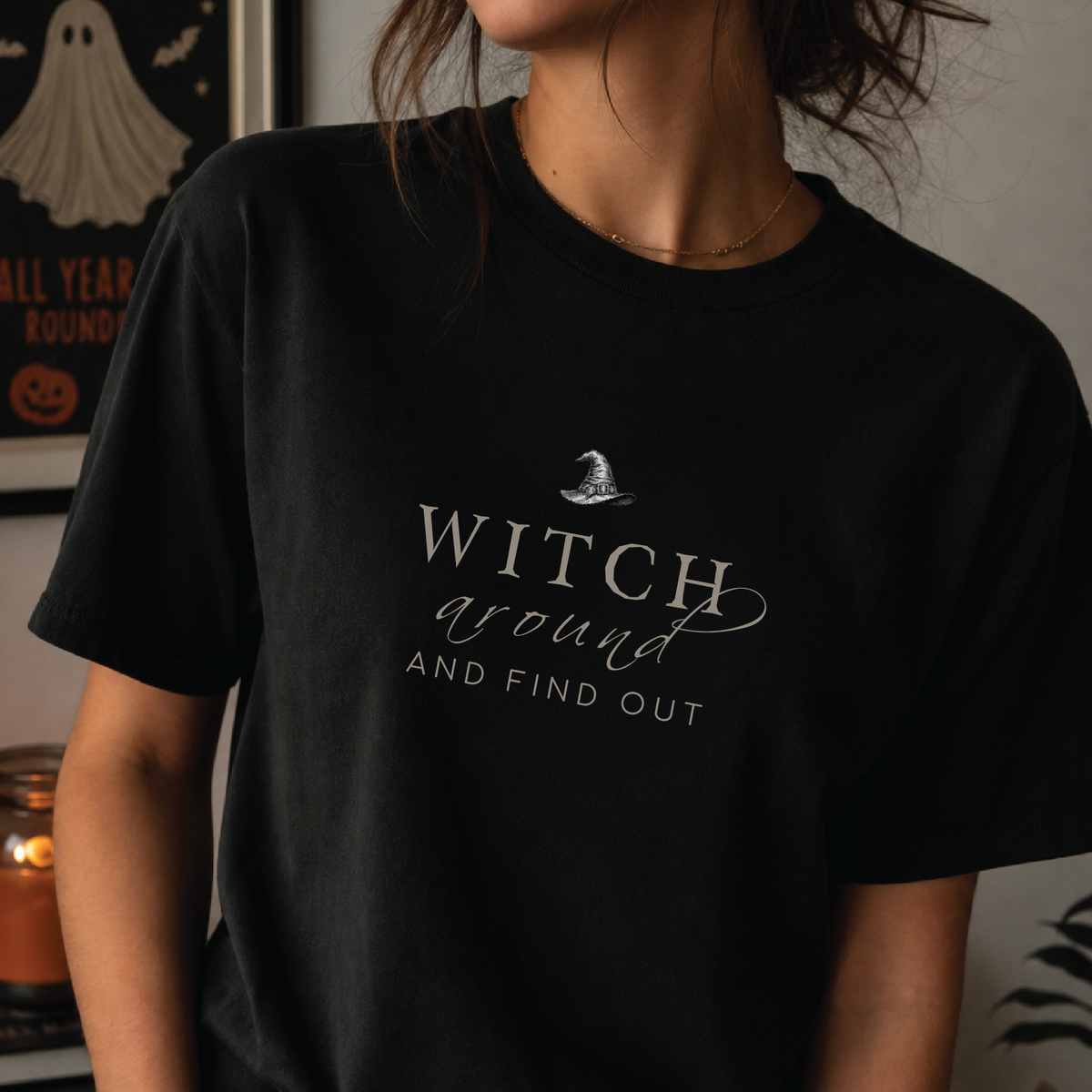 A person wearing a black short-sleeve graphic T-shirt with the phrase 'WITCH around AND FIND OUT' in white script and a small witch hat illustration above the text. The photo is taken indoors with a softly blurred Halloween-themed wall and candle in the background.